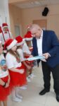 Mężczyzna wręcza czekoladki dziecku. Dzieci ubrane są w biało czerwone stroje, na głowie mają świąteczne czapki św. Mikołaja.