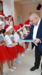 Mężczyzna wręcza czekoladki dziecku. Dzieci ubrane są w biało czerwone stroje, na głowie mają świąteczne czapki św. Mikołaja.