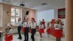 Dzieci ubrane w biało czerwone stroje z czapkami mikołaja. Tańczą i śpiewają.
